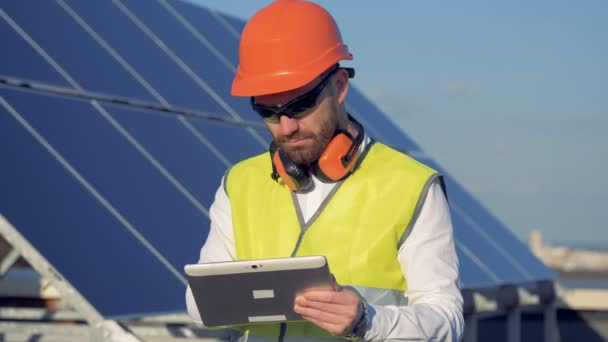 Engineer uses his tablet to work, standing next to solar panels on the roof. - Filmati, video