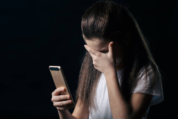 Μία θλιβερή έφηβος κρατώντας ένα κινητό τηλέφωνο, θρηνώντας κάθεται στο κρεβάτι στην κρεβατοκάμαρά της με ένα σκούρο φως στο παρασκήνιο - Φωτογραφία, εικόνα