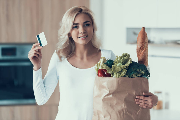 Blonde woman with credit card and package of products is standing in kitchen. - Foto, immagini