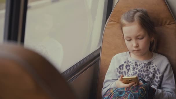 Joven, hermosa pasajera con bolso escolar en el autobús escolar en movimiento utilizando la red social en su teléfono inteligente y mirando por la ventana
. - Imágenes, Vídeo