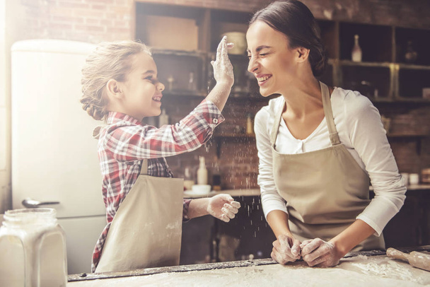 Beautiful young mom and her cute little daughter are playing and smiling while baking in kitchen at home - Фото, изображение