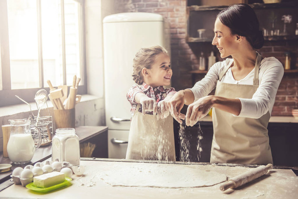 Cute little girl and her beautiful mother are sprinkling the dough with flour and smiling while baking - Foto, imagen
