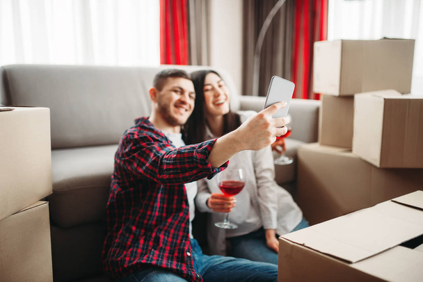 love couple sitting on the floor near cardboard boxes, taking selfie and celebrating  moving to new house - Fotoğraf, Görsel