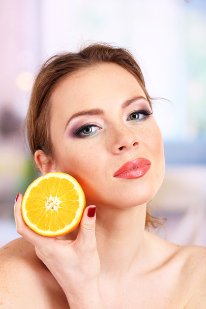 Beautiful young woman with bright make-up, holding orange, on bright background - Photo, Image