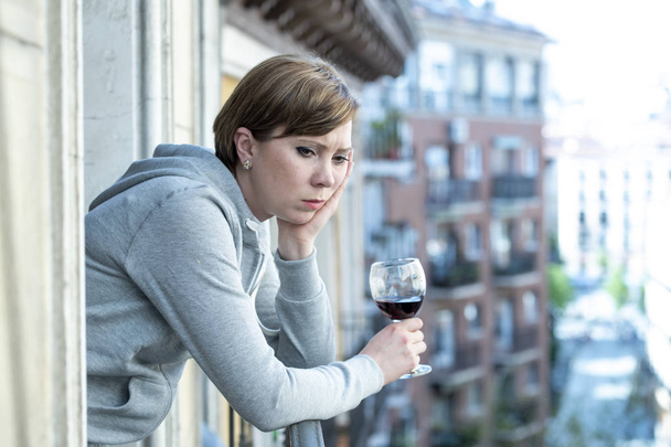 Beautiful red haired caucasian woman suffering from depression holding a glass of wine on a balcony at home. Staring out feeling sad, pain and grief. Crisis, depression and mental health concept - Foto, Imagem