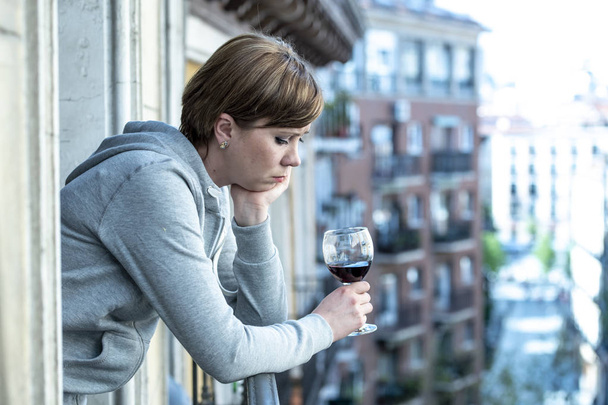 Beautiful red haired caucasian woman suffering from depression holding a glass of wine on a balcony at home. Staring out feeling sad, pain and grief. Crisis, depression and mental health concept - Foto, Imagem