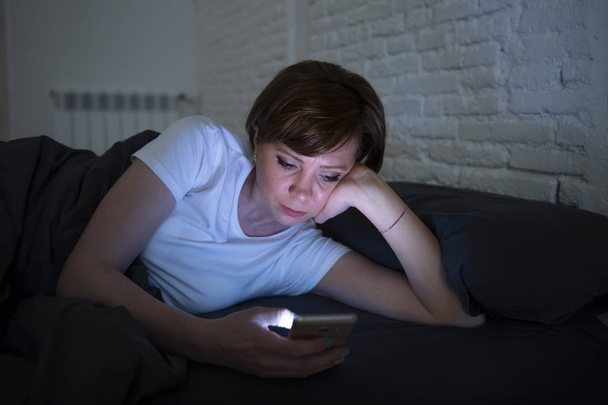 Young attractive woman awake late at night using smart phone lying in bed in a dark bedroom. Using mobile for chatting and sending messages in internet addiction, mobile abuse and insomnia concept - Valokuva, kuva