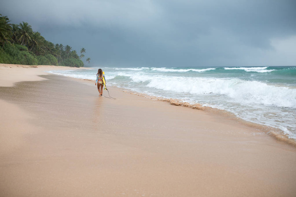 Back view of a attractive woman holding surf board in hands on coastline. - Foto, afbeelding