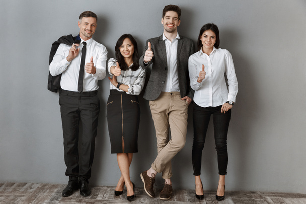 multicultural business people in formal wear showing thumbs up while standing at grey wall - Photo, Image