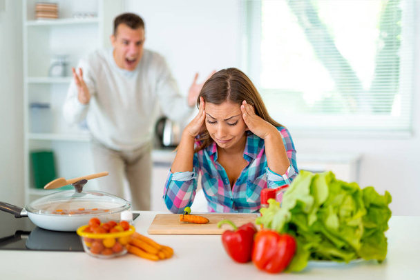 Young couple arguing in the domestic kitchen. Upset woman sitting and holding head. Angry man shouting and waving arms. Selective focus. - Photo, Image