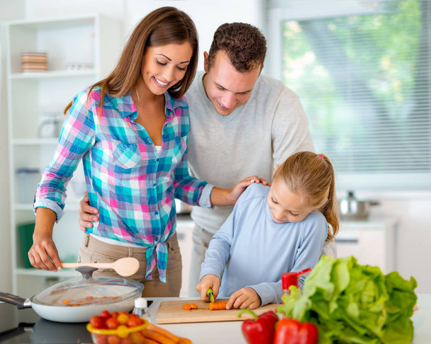 Kaunis nuori perhe ruoanlaitto terveellisen aterian kotitalouskeittiössä. Pikkutyttö oppii leikkaamaan porkkanaa keittiön hallituksessa ja hänen vanhempansa valvovat häntä.
. - Valokuva, kuva