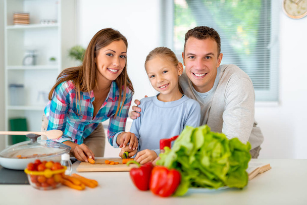 Krásná mladá rodina vaření zdravé jídlo v domácí kuchyni. Malá holčička se učí, jak snížit mrkev na kuchyňské desce a její rodiče dohlížet na ni. Při pohledu na fotoaparát. - Fotografie, Obrázek