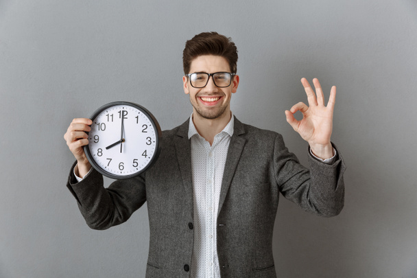 portrait of smiling businessman with clock in hand showing ok sign on grey wall background - Photo, Image