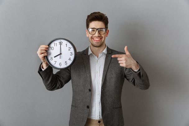 portrait of smiling businessman pointing at clock in hand and looking at camera on grey wall background - Фото, изображение