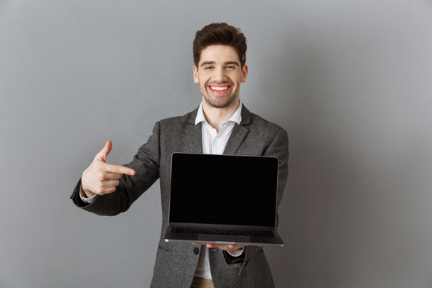 portrait of smiling businessman in suit pointing at laptop with blank screen against grey wall background - Fotó, kép