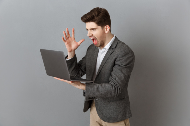 frustrated businessman in suit with laptop against grey wall background - Photo, Image