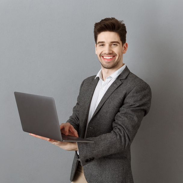 sonriente hombre de negocios en traje con portátil contra fondo de pared gris
 - Foto, Imagen