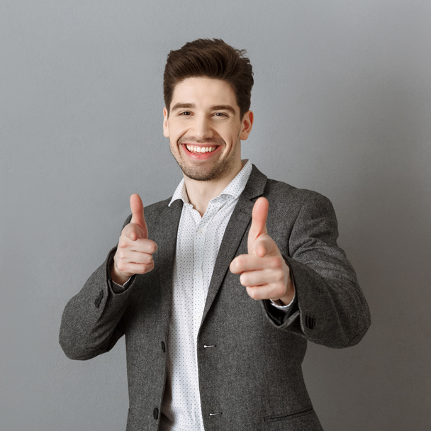 muotokuva hymyilevä liikemies puku osoittaa kameraa vastaan harmaa seinä
 - Valokuva, kuva