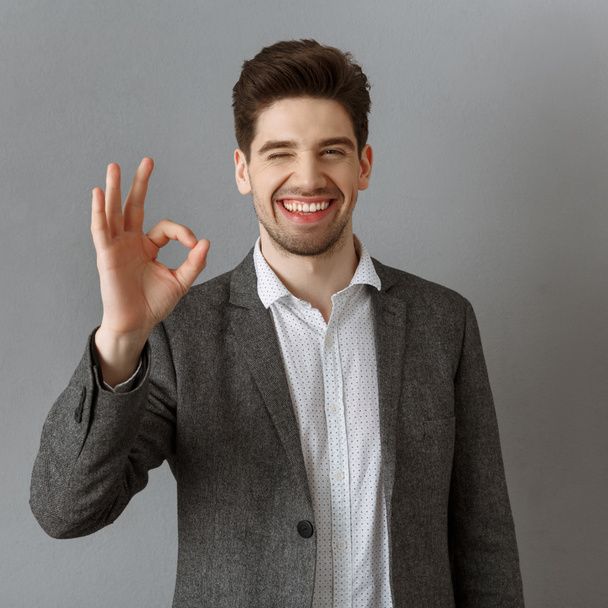 muotokuva hymyilevä liikemies puku osoittaa ok merkki vastaan harmaa seinä
 - Valokuva, kuva
