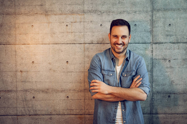 Młody przedsiębiorca udany uśmiechający się stojąc przed betonową ścianę z skrzyżowane ręce i patrząc na kamery. - Zdjęcie, obraz