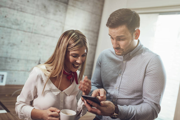 Молодые улыбающиеся предприниматели делают перерыв на кофе и пользуются смартфоном в офисе
. - Фото, изображение