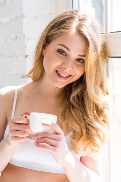 beautiful cheerful girl with cup of coffee standing near window  - Foto, Imagem
