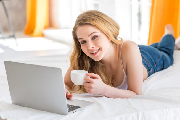 attractive girl with cup of coffee using laptop on bed - Photo, Image