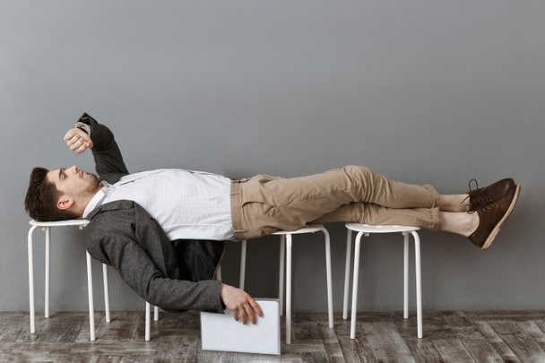 tired businessman with documents checking time while lying on chairs and waiting for job interview - Photo, Image