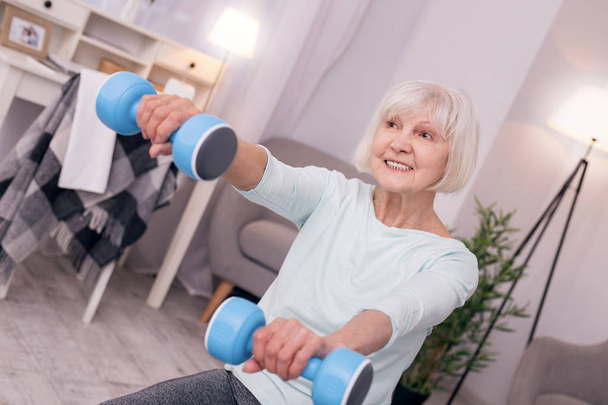 Pleasant elderly woman holding blue dumbbells and smiling - Photo, Image