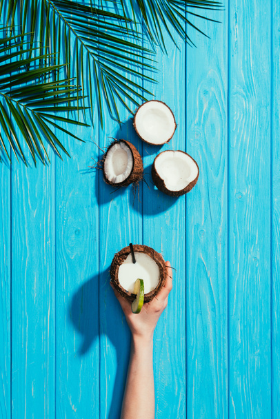 top view of human hand with coconut cocktail, cracked coconuts and palm leaves on turquoise wooden surface - Photo, Image