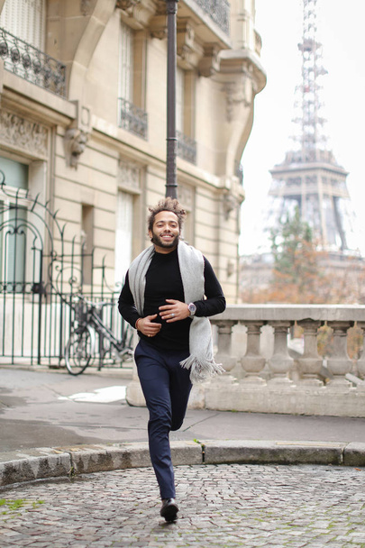 Afroamerikaner läuft mit Eiffelturm im Hintergrund, trägt grauen Schal und schwarzen Pullover. - Foto, Bild