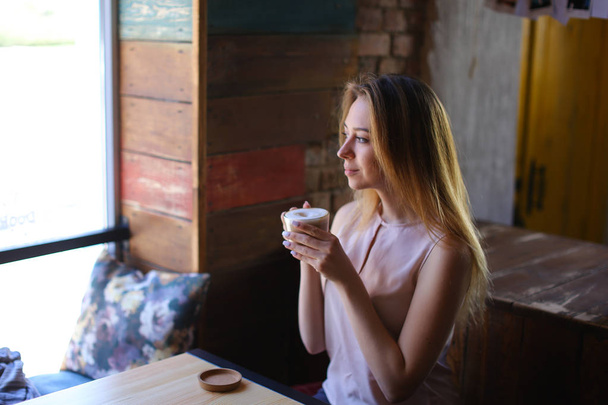 Chica bonita sentada cerca de la mesa con una taza de café y sonriendo
. - Foto, Imagen