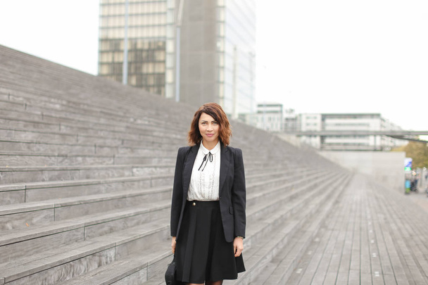 Glückliche Geschäftsfrau steht auf Treppen mit Tasche und hohen Gebäuden im Hintergrund. - Foto, Bild