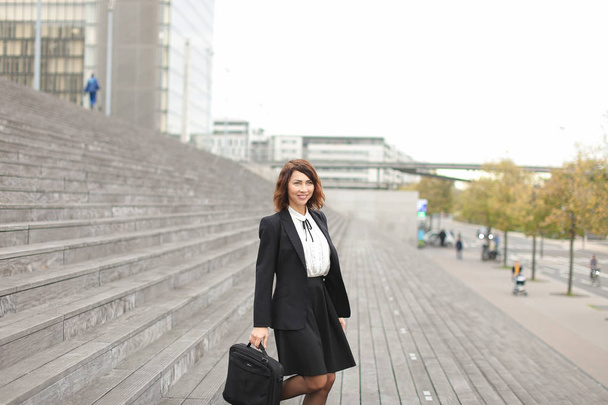 Θηλυκό Γραμματέας στέκεται στις σκάλες με τσάντα και ψηλά κτίρια στο φόντο. - Φωτογραφία, εικόνα