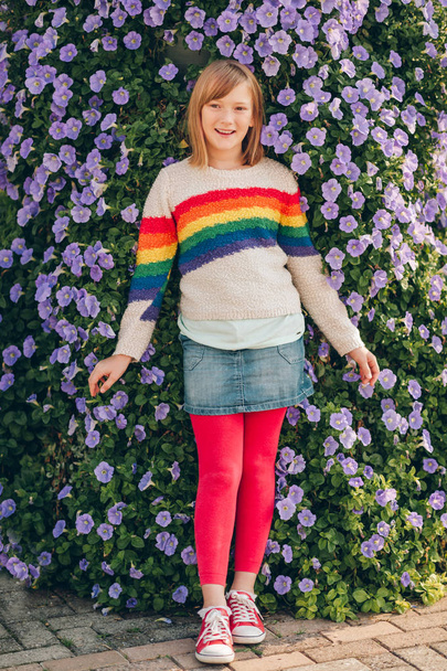 朝顔の花の壁の横にポーズ虹のプルオーバーとデニムのスカートをはいているかわいい女の子の屋外のポートレート - 写真・画像