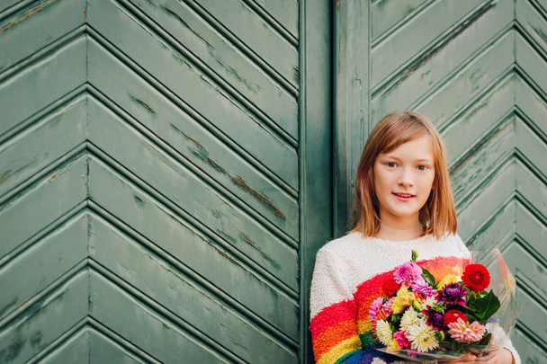 Outdoor portrait of pretty kid girl wearing colorful pullover, holding bouquet of autumn flowers, posing against old green vintage door - Photo, image