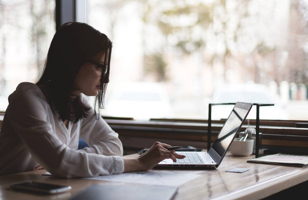 Брюнетка Деловая женщина в очках работает за ноутбуком и с бумагами в уютной атмосфере кафе. Силуэт фото на фоне окна
 - Фото, изображение