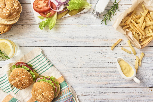 Preparation of fresh tasty burgers. Green raw vegetables on cutting board and buns in plate, french fries and class with lemonade on white wooden background. Top view. Space for text - Photo, Image