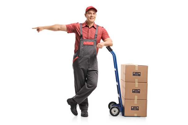 Full length portrait of a mover leaning on a hand truck with a stack of boxes and pointing isolated on white background - Fotoğraf, Görsel