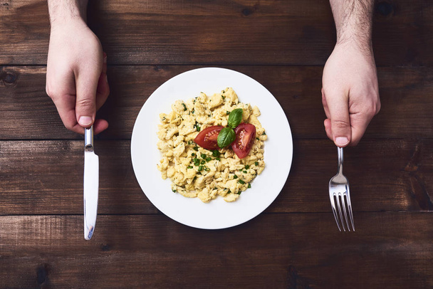 Man preparing to eating tasty scrambled egg with tomato and basil in white plate over dark brown rustic wooden table background. Top view - Φωτογραφία, εικόνα