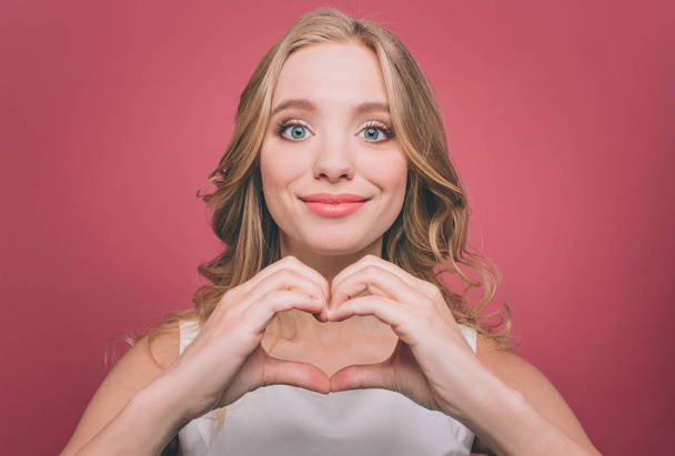 Lovely and pretty girl is making a heart from her fingers. She is looking on camera with grateful sight. She is smiling. Isolated on pink background. - Foto, immagini