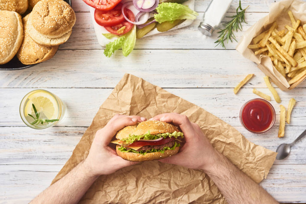 Homme se préparant à manger des hamburgers savoureux frais. Légumes crus verts sur planche à découper et petits pains dans une assiette, frites et classe avec du cola sur fond blanc en bois. Vue du dessus
 - Photo, image