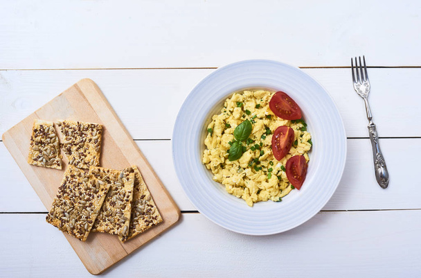 Tasty Scrambled egg with basil and tomato in white plate and fork. Crispbread on cutting board over white wooden background. top view - Fotó, kép