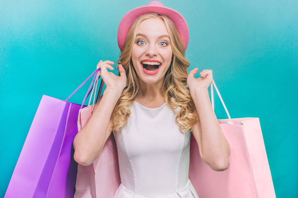 Cheerful and attractive girl is holding colorful bags behind her back. Also she wears pink hat. Isolated on blue background. - Fotografie, Obrázek