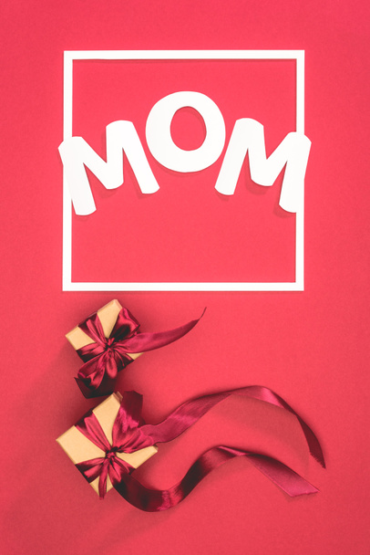 top view of gift boxes with ribbons and word mom in frame on red surface, mothers day concept - Φωτογραφία, εικόνα