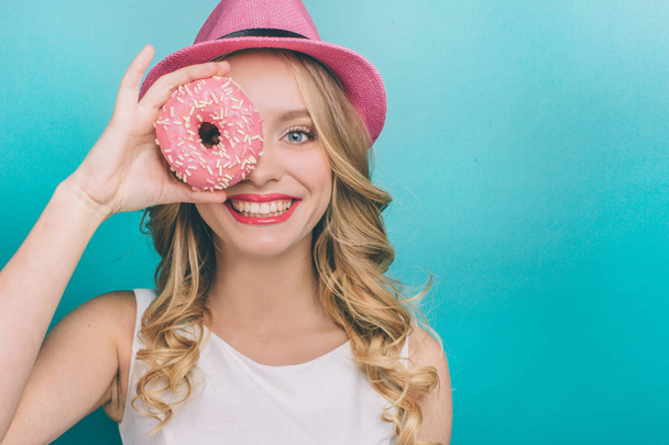 Attractive girl with blonde hair is looking straight and smiling. She is covering one of her eyes with donut with pink glaze. Girl is happy. Isolated on blue background. - Foto, Bild