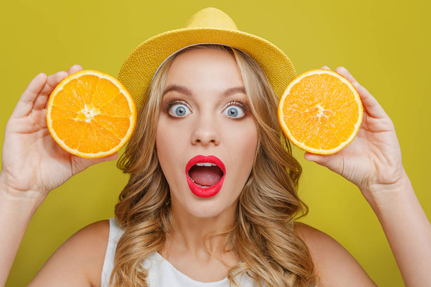 Amazed and excited young woman wears yellow hat. Also she is showing two pieces of juicy orange to camera. She is keeping her mouth wide open. Isolated on yellow background. - Foto, Imagem