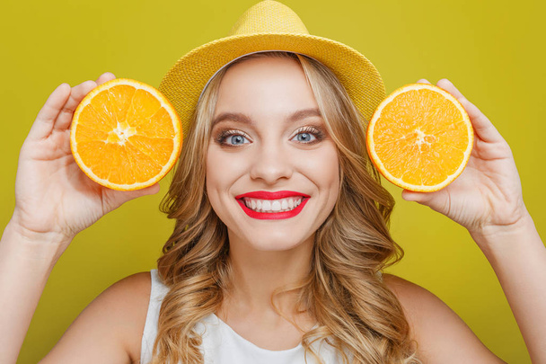 Beautiful girl is holding juicy pieces of orange and showing it to camera. She is looking straight forward and smiling with a cheerful smile. Isolated on yellow background. - Foto, afbeelding
