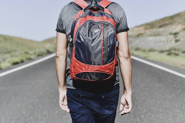 closeup of a young caucasian man, seen from behind, carrying a backpack walking by a secondary road - Photo, Image