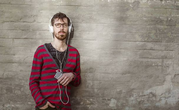 Młody człowiek słucha muzyki ze smartfona. Hipster z brodą w okularach w słuchawki z telefonem na tle ceglany mur. - Zdjęcie, obraz
